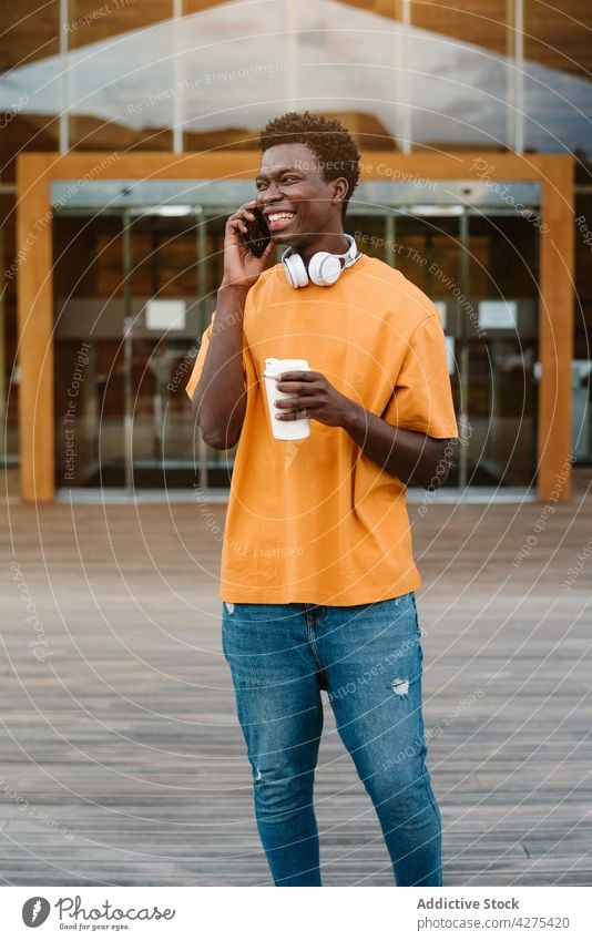 Cheerful black man with cup of coffee talking on smartphone speak laugh using headphones to go beverage conversation male device cellphone connection