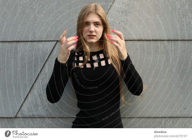 Young woman in black dress in front of grey metal wall raises her hands towards camera to fix her long blond hair, showing pink painted fingernails Woman Dress