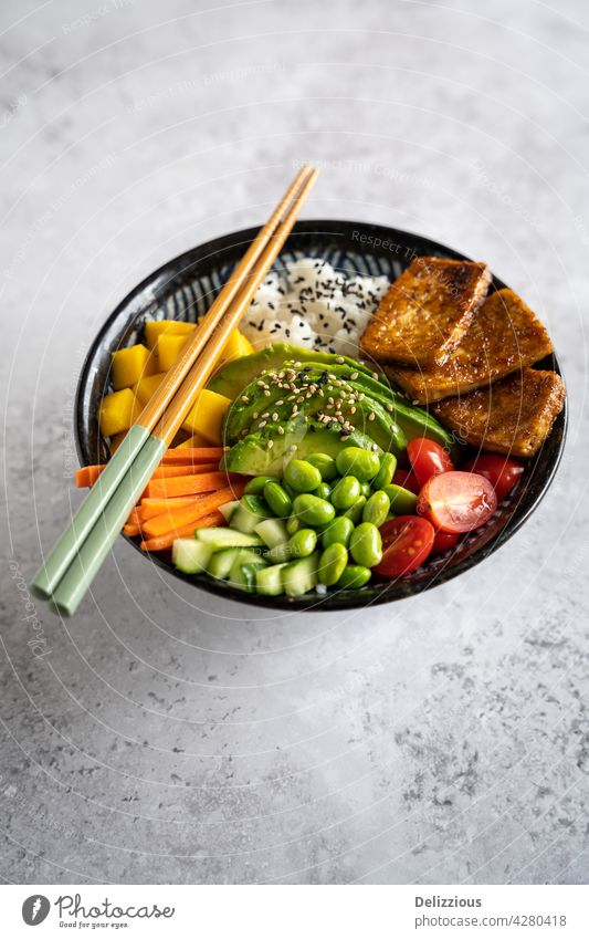 Side down view of a homemade vegan poke bowl with tofu, mango, avocado, carrot, edamame, tomato, cucumber on a grey background, vertical asian chopsticks