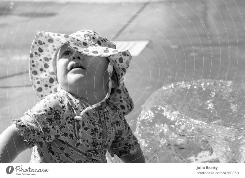One year old baby at a splash pad water park on a hot day; toddler wears sun protective UPF sun hat and swimsuit as she looks up at parents for reassurance