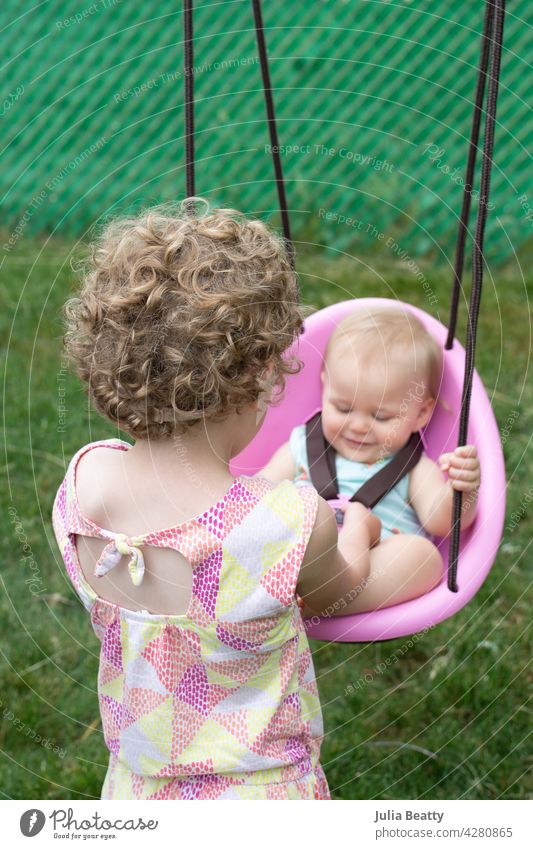 Five year old girl helps one year old cousin in pink swing; baby smiling sisters cousins bucket swing play set gym backyard garden curly pixie girls female