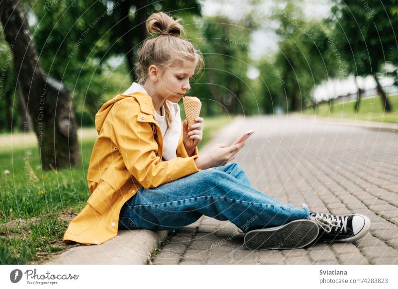 A blond girl sitting on the road with a phone and ice cream communicates in social networks. Child outdoors summer park teenager smartphone selfie social media