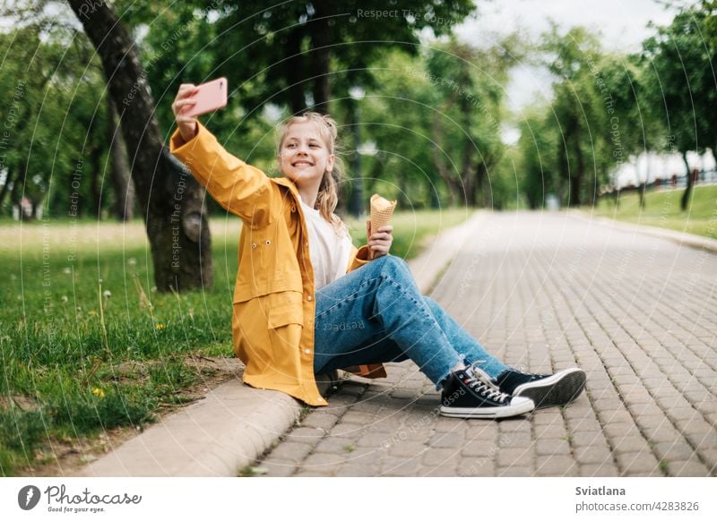 A blonde sitting on the road with a phone and ice cream makes a funny selfie. Child outdoors summer park teenager smartphone social media trend happy cute child