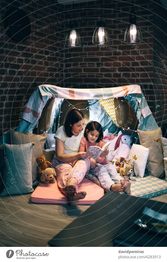 Mother and daughter having a pajama party reading a book mother telling diy tent teepee together cozy enjoyment child family home camping caucasian love girl
