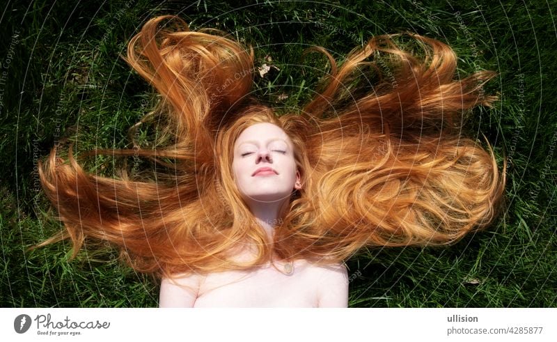 Top view to beautiful portrait of young sexy redhead woman, lying contend in the spring, summer sun, relaxing on the green grass, the long red hair draped freely around the head, copy space