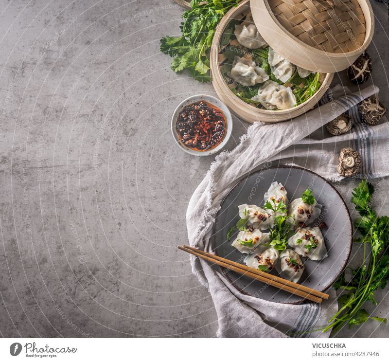 Asian food concept with homemade dumplings on plate and typical equipment and ingredients like chopsticks, steamer, fresh asian herbs and chili sauce. Dark concrete background. Top view. Food border