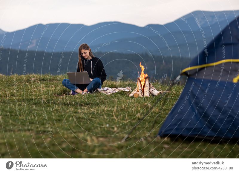 young female freelancer working on laptop in the mountains in the evening. Tourist girl sitting near campfire and tent. Copy space tourist vacation