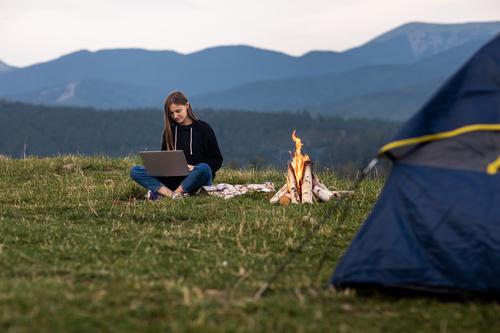 young female freelancer working on laptop in the mountains in the evening. Tourist girl sitting near campfire and tent. Copy space tourist vacation