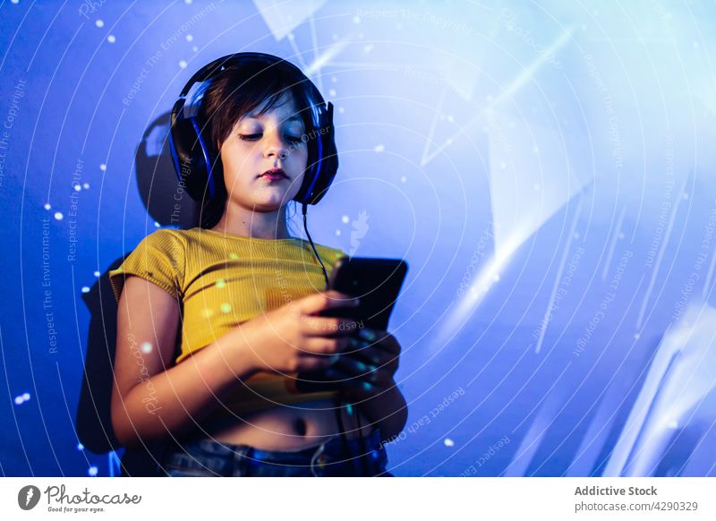 Little girl using tablet and listening to music in headphones browsing gadget neon light kid casual surfing connection enjoy device modern unemotional child