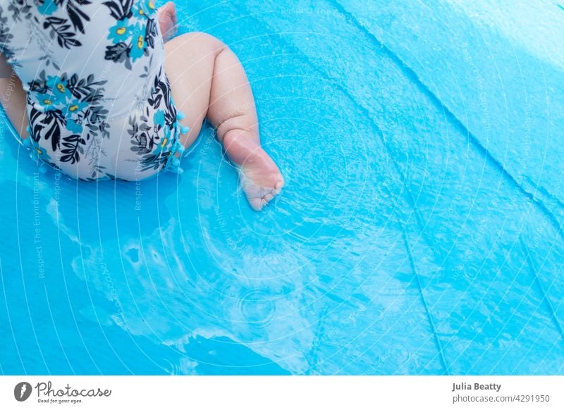 Young baby sitting in a blue inflatable swimming pool; toddler wearing a floral swimsuit water safety sun safety blow up pool kiddie pool hands and knees seated