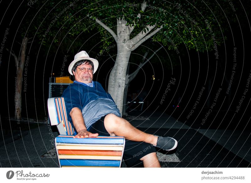 " He's pissed " middle aged man, sitting on a colorful park bench in the evening casually wearing a white hat and glasses , shorts and sandals and contorting his face.