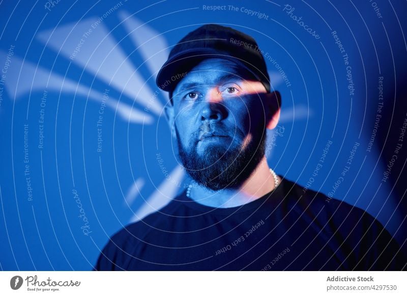 Serious bearded male in dark neon studio man serious room alone shadow appearance confident individuality adult light casual cap illuminate pensive brutal