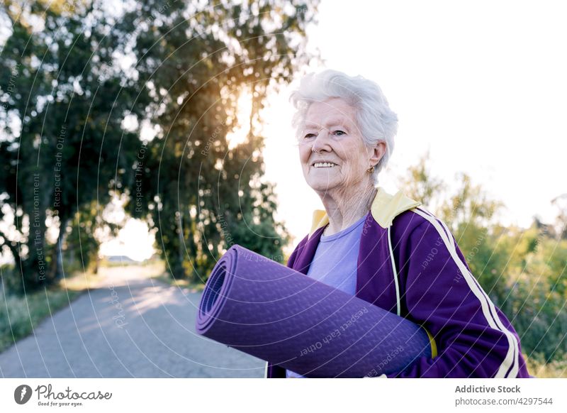 Happy elderly sportswoman with mat in countryside training fitness morning smile carry healthy sportswear female aged senior wellness lifestyle athlete workout