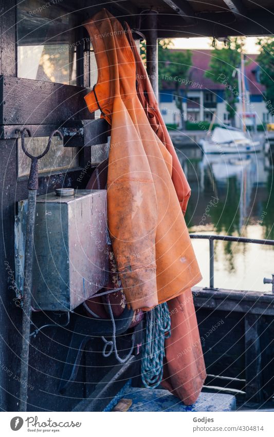 Frisian mink, hoe, rope hanging on a fishing boat. In the background a sailboat on the water and a house Raincoat Rain pants Clothing Hoe friesennerz Orange