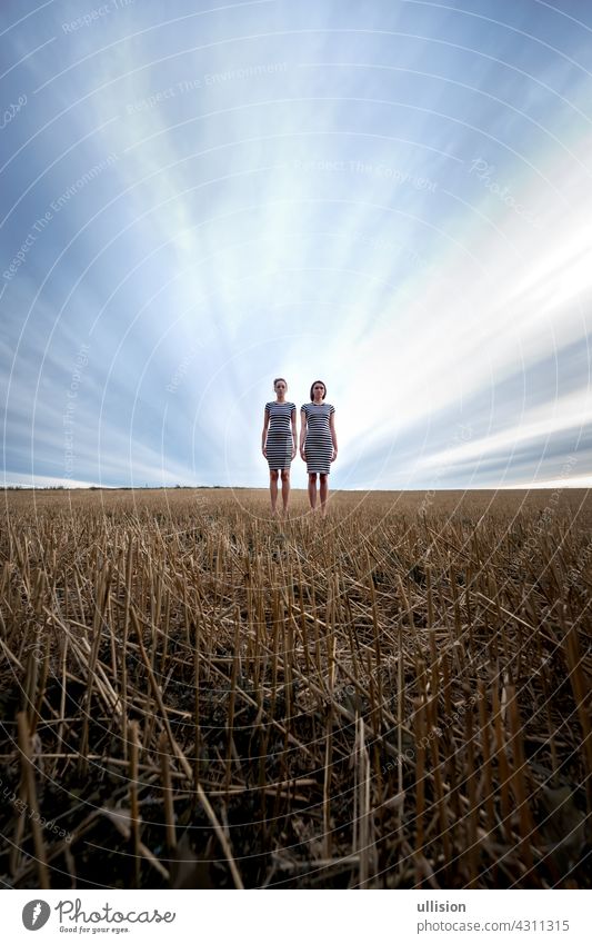 Two ghostly looking, eerie young women in striped dresses stand against dramatic sky in a stubble field, wide angle, copy space. Hallowe'en Mystery Woman spirit