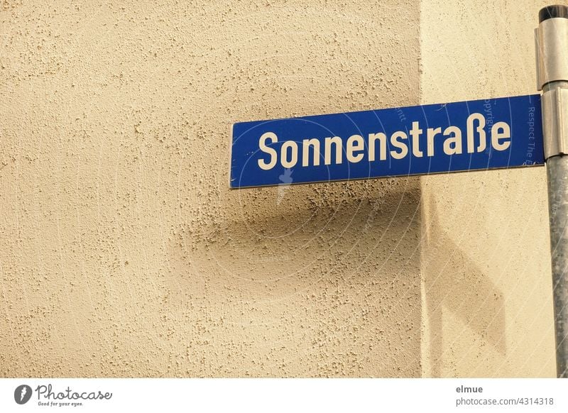 blue street name sign with white writing " Sonnenstraße " on a metal pole in front of a house corner with shadow / live / navigation Street sign Sun Street