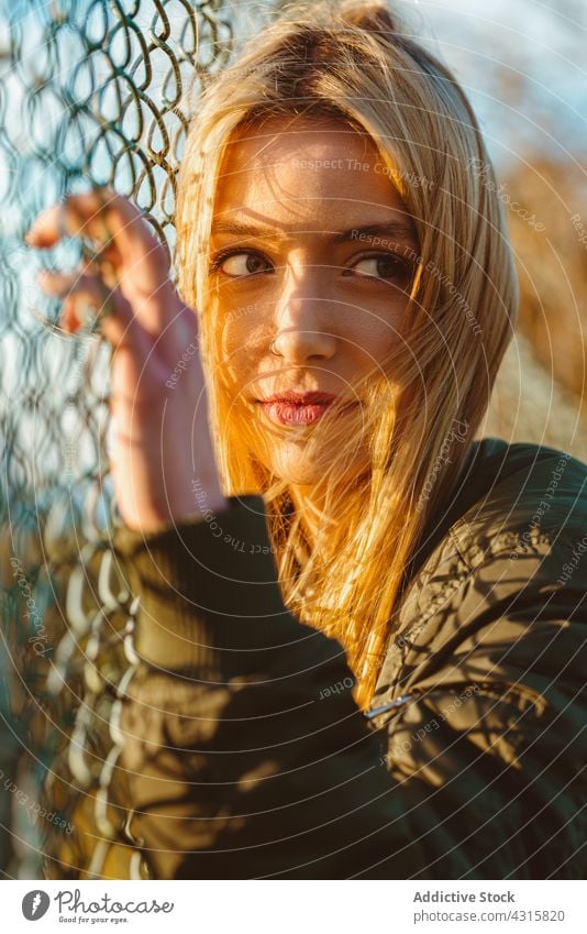 Calm woman leaning on net fence in sunlight portrait chain link calm blond charming modern beauty young bright carefree alone jacket long hair style natural