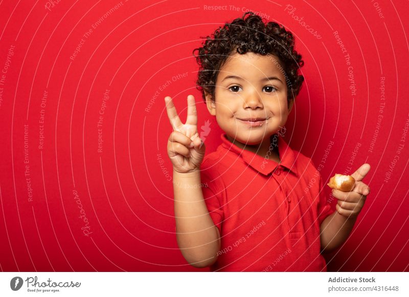 Content boy with donut showing two fingers gesture child v sign eat doughnut victory kid tasty delicious sweet childhood cheerful studio joy happy smile dessert