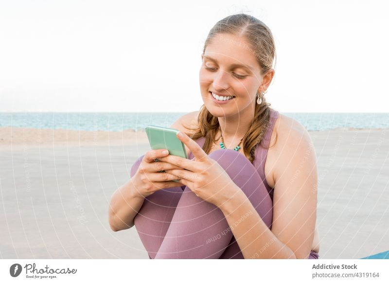 Smiling woman on smartphone on yoga mat mobile yogi beach smile female gadget browsing cheerful using happy slim text message device sit enjoy wellness content