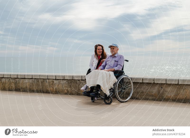 Cheerful adult daughter and aged father in wheelchair on promenade man senior chill sea embankment together seashore disable handicap elderly happy smile