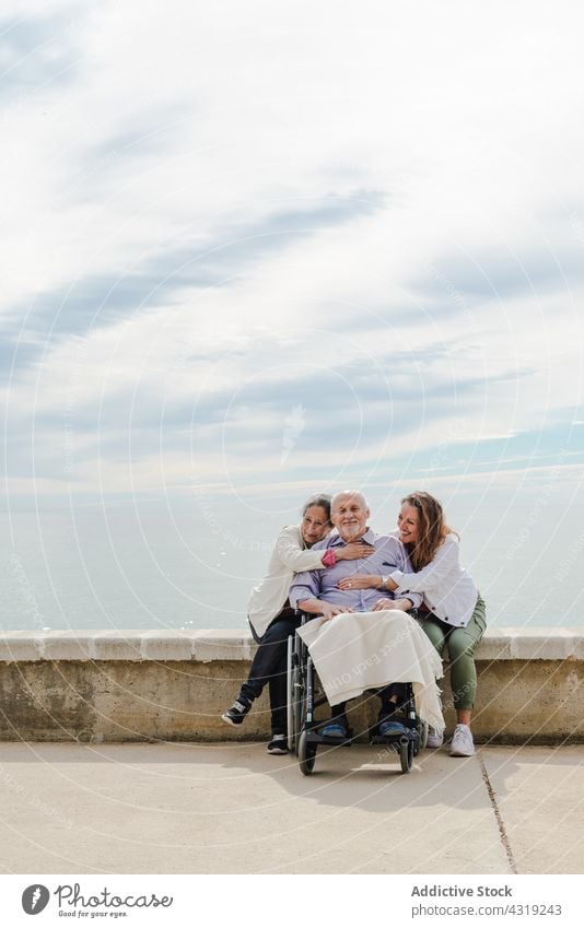 Cheerful family embracing father in wheelchair senior promenade hug unity chill embankment together sea seashore adult disable handicap embrace cuddle elderly