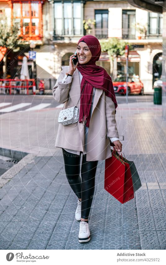 Smiling Muslim woman with shopping bags in city talk smartphone paper bag shopper mobile call hijab female muslim ethnic gadget speak walk shopaholic using