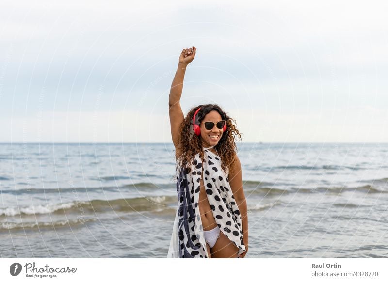 Happy young beautiful afro american woman smiling , wearing headphones dancing and raising arm on the beach.Female enjoying summer holidays concept lifestyle.