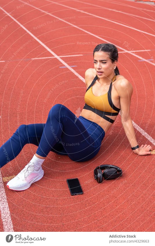 Athletic woman taking break on stadium track sportswoman athlete rest smartphone headphones workout tired fitness training young recreation sneakers ethnic