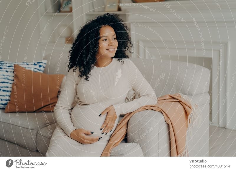 Beautiful young mixed race pregnant woman hugging her tummy sitting pregnancy african sofa relax rest think dream happy baby love child abdomen maternity mom