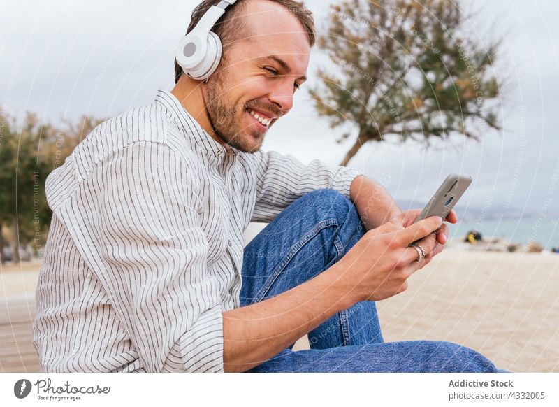 Happy man in headphones using smartphone on beach happy listen gadget music male cheerful young style summer wireless device enjoy lifestyle sea browsing mobile