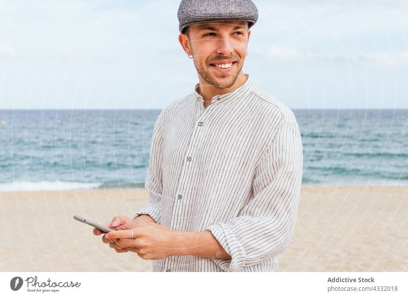 Cheerful man with smartphone on sandy beach using style happy sea trendy smile male young beard look outfit fashion positive cheerful browsing mobile gadget