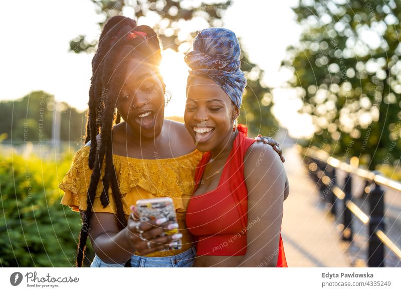 Two female friends with smartphone, smiling outdoors diversity millennials together friendship sisterhood black live matter afro proud real people candid