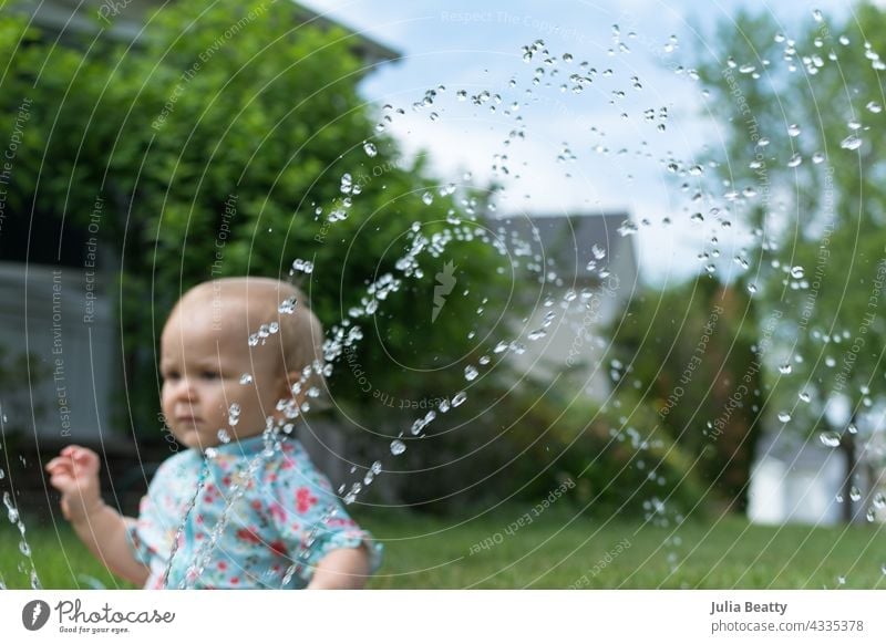 Toddler seated in grass in front yard; in front of her a sprinkler sprays water baby toddler sun safety water play home neighborhood midwest spf upf