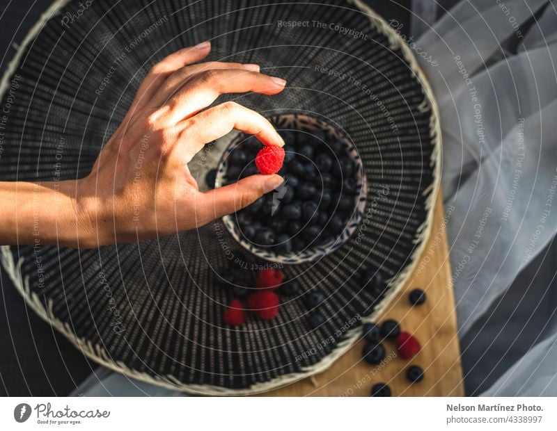 Hand holding a raspberry with a plate and blueberries in the background snack closeup handful organic grocery dieting woman food antioxidant heap nature people