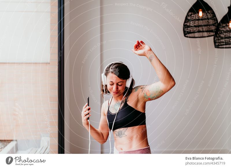 happy muscular caucasian woman listening to music on mobile phone and headset in the gym. Dancing by window during daytime. Sport and healthy lifestyle