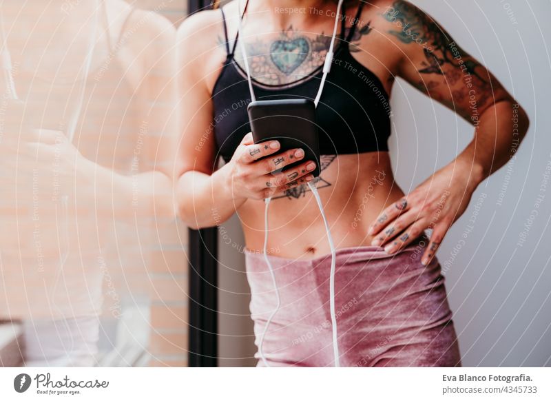 unrecognizable muscular caucasian woman listening to music on mobile phone and headset in the gym. Standing by window during daytime. Sport and healthy lifestyle