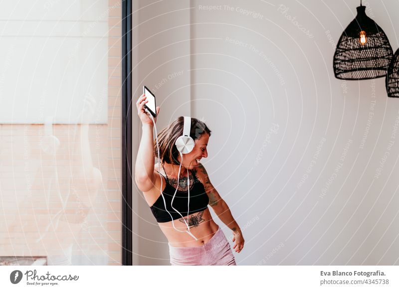 happy muscular caucasian woman listening to music on mobile phone and headset in the gym. Dancing by window during daytime. Sport and healthy lifestyle