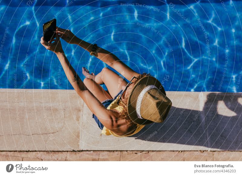 top view of young caucasian woman sitting by pool side using mobile phone to take a selfie. Summer time, vacation and lifestyle technology swimming pool