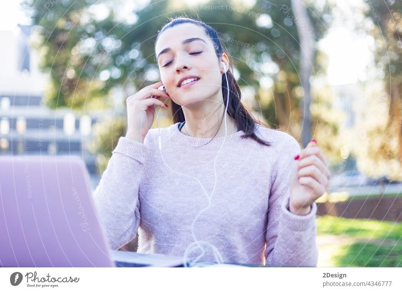 Image of feminine young woman 20s in casual wear relaxing in green park, and using mobile phone with earphones girl wireless music sitting female outdoors