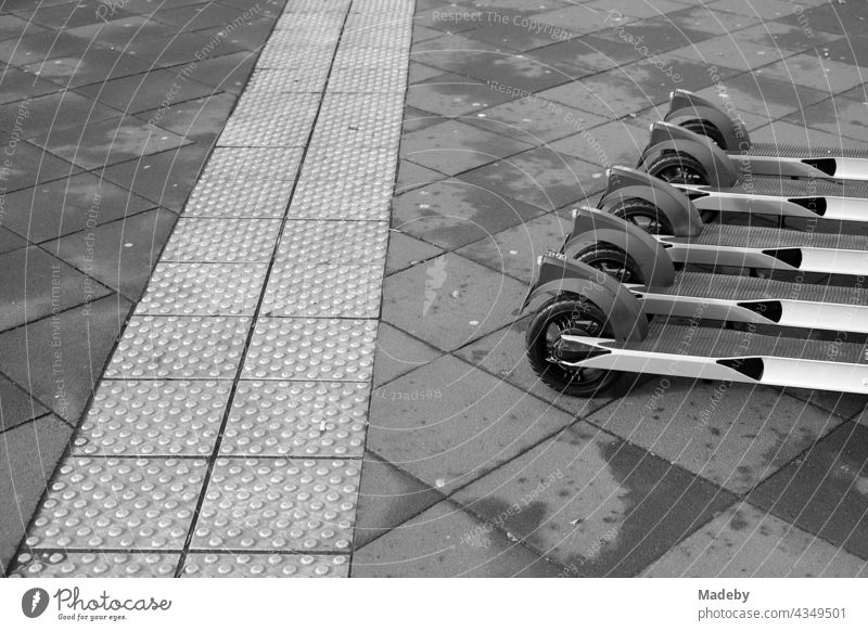 Modern e-scooters lined up for rent next to the tactile guidance system for the visually impaired on the pavement in the city centre of Frankfurt am Main in Hesse, photographed in neo-realistic black and white