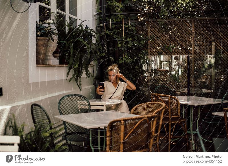 Blonde woman taking a selfie in the terrace of a restaurant confortable Lifestyle portrait smartphone Self-confident tables isolated alone blonde caucasian