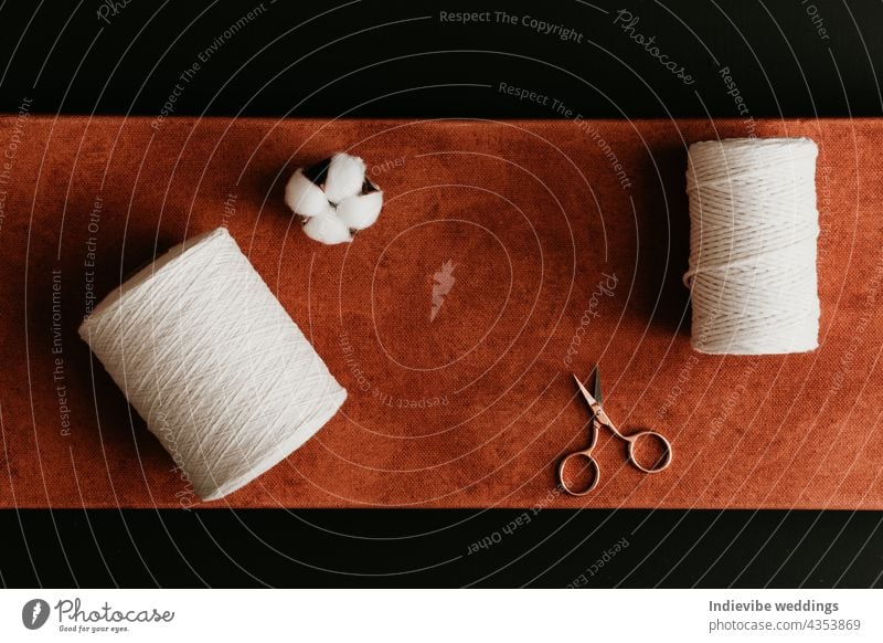 Two rolls of cotton rope, a gold rose scissors and a white cotton flower head on rust and orange color canvas on black background. brown string thread creative
