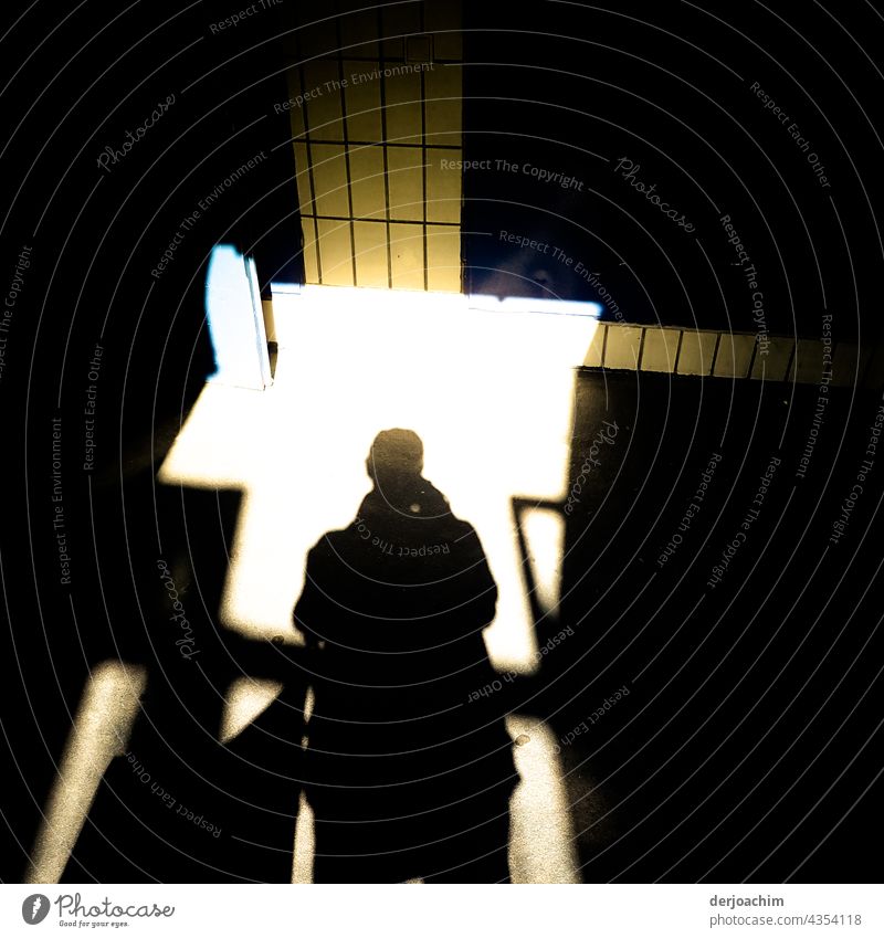 Der Fotograf als schwarzer großer Schatten,  tritt  in das Treppenhaus das vor ihm von hellem Sonnenlicht durchflutet ist. Vor ihm und rechte sind an der Wand gelbe Fliesen angebracht.