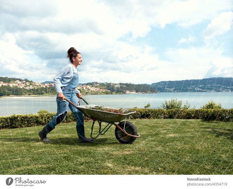 Teenage girl pushing a wheelbarrow in the garden farmer woman agricultural tool green grass outdoors outside person one single action working agriculture female