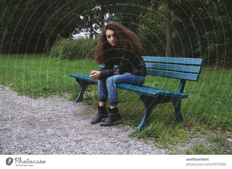 Young woman sitting on park bench teenager Sit Park bench Wait Brunette Curl Jeans somber Loneliness on one's own Looking into the camera Youth (Young adults)