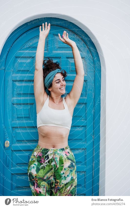 portrait of happy caucasian woman with arms raised in front of blue door during summer time. Outdoors lifestyle outdoors city beach carefree smiling smile town