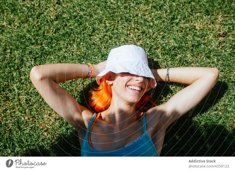Cheerful woman lying on grass summer sunny happy park green beautiful redhead hat nature young happiness person relax female spring field lawn people meadow