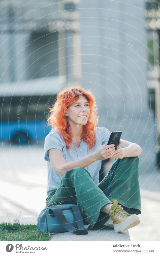 Delighted woman with red hair listening to music in earphones in street smartphone browsing redhead message social media song female using smile device internet
