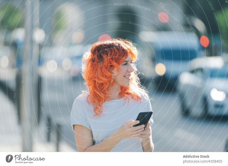 Delighted woman with red hair listening to music in earphones in street smartphone browsing redhead message social media female using smile device internet