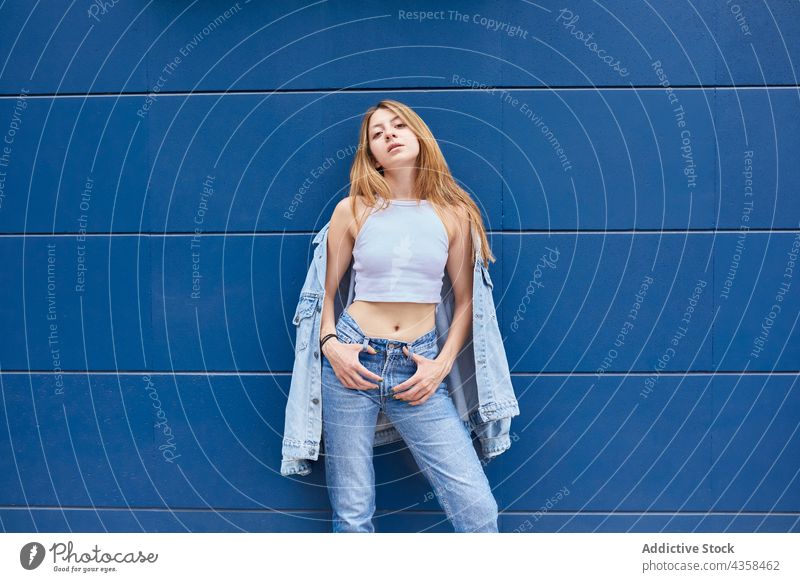 Confident stylish woman in denim outfit standing against blue wall style jeans city cool trendy fashion jacket street female urban confident serious appearance